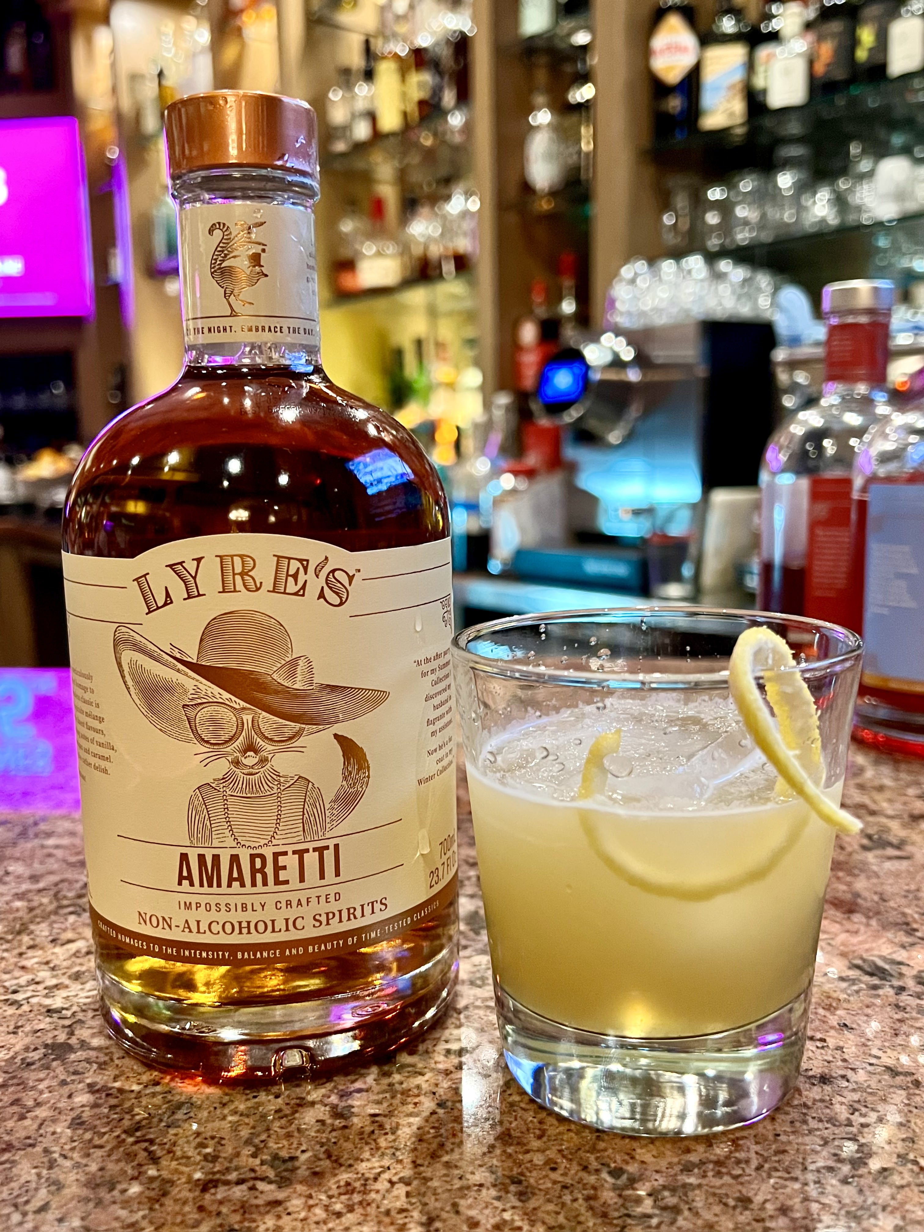 Geateano's Amoretti Sour mocktail - Lyre's Amaretto non-alcoholic spirits bottle with cat wearing hat and glasses logo beside glass with lemonade colored drink topped with a lemon twist